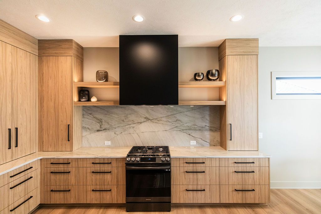 Modern kitchen features light wood cabinetry with sleek black hardware and a black range hood. A marble backsplash and countertops add elegance.