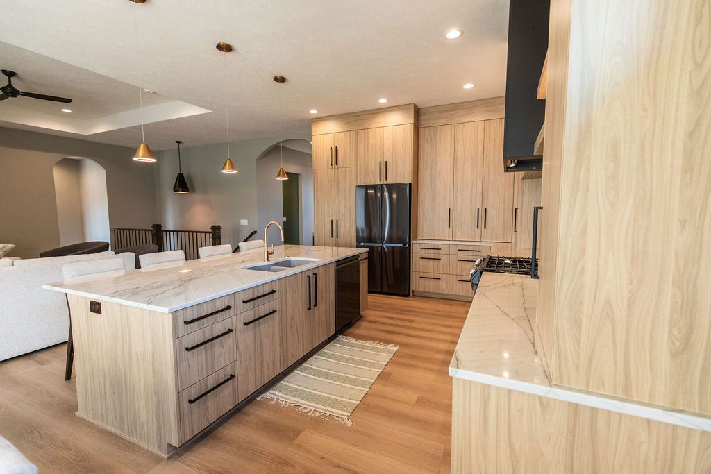 Modern kitchen features a spacious island with marble countertops and light wood cabinets with black hardware. Stainless steel appliances, including a refrigerator and dishwasher, are present alongside a light-colored wood floor and stylish pendant lighting.