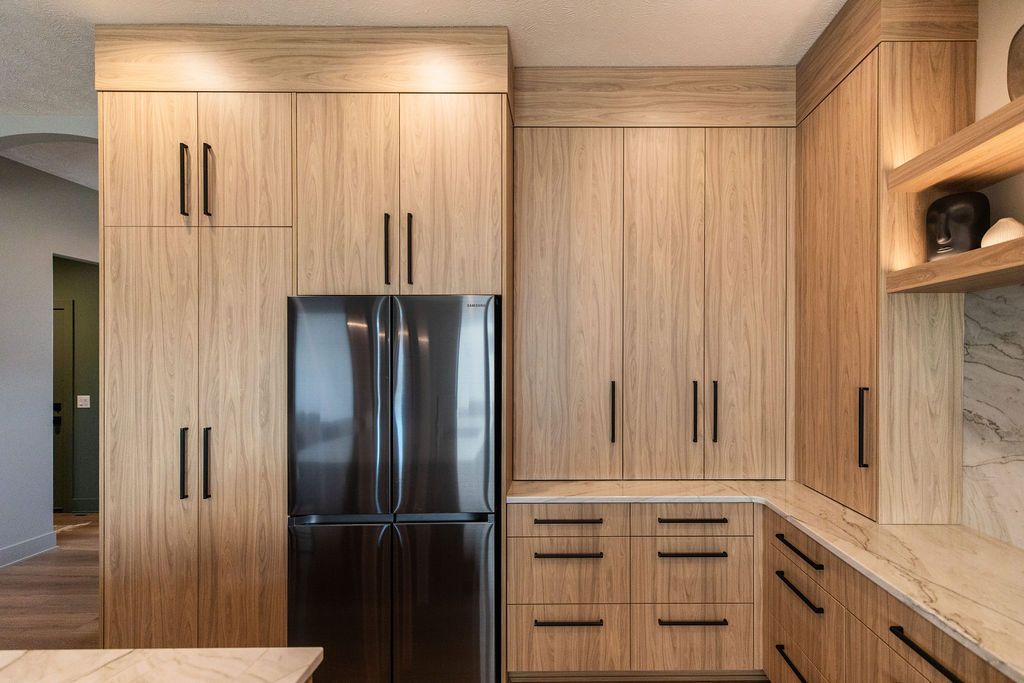 Modern kitchen with light wood cabinetry and black hardware, featuring a stainless steel refrigerator and marble countertops. Recessed lighting illuminates the clean, minimalist design.