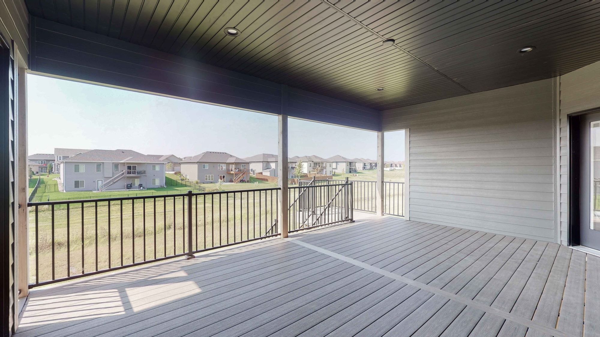 Covered deck featuring gray wood decking and a black metal railing overlooks a grassy yard with houses in the distance. The ceiling is a dark gray and has recessed lighting.