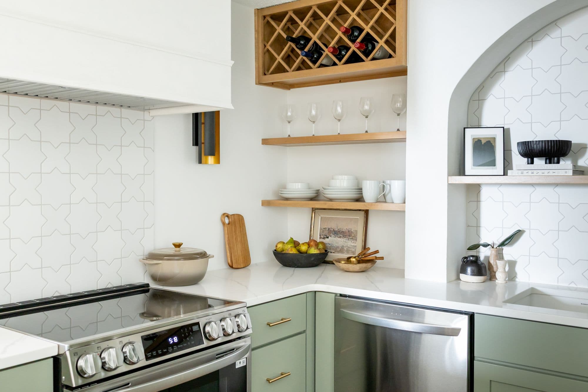 Modern kitchen area features light green cabinets with stainless steel appliances and white countertops. An archway and white tile backsplash complement the warm wood accents and decor.